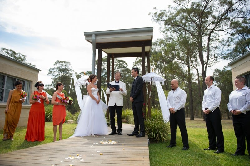 Bride and groom under a wooden pergola at a private house in the Hunter Valley NSW with celebrant and bridesmaids watching
