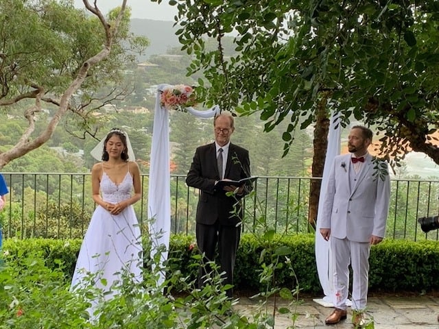 Bride and groom with celebrant under a ceremony arch at Bible Garden, Palm Beach NSW